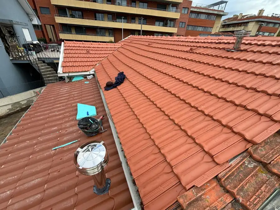 Pose de tuiles en cours : grand pan de toiture fraîchement recouvert de tuiles rouges neuves, arêtier et faîtage posés ; au premier plan un seau d'outils, un marteau.