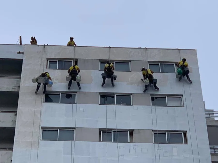 Ravalement de façade en cordiste : cinq ouvriers en tenue jaune suspendus en rappel le long de la façade d'un immeuble, seaux d'enduit/peinture à la main.